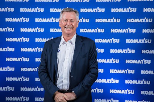 Man wearing suit standing in front of blue wall with Komatsu logo